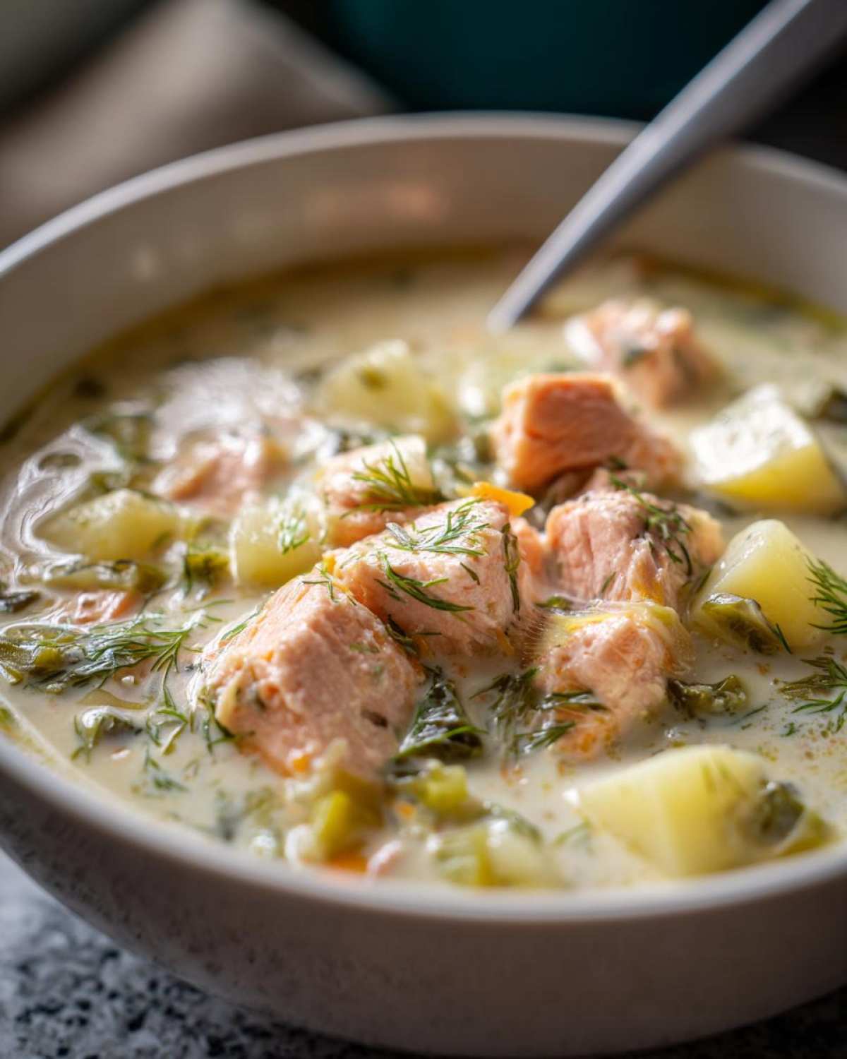 Close-up of a bowl of creamy salmon soup featuring chunks of pink salmon, potatoes, greens, and fresh dill.