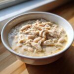 A close-up of a white bowl filled with creamy white chicken chili, featuring shredded chicken and white beans.