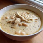 A close-up of a white bowl filled with creamy white chicken chili, featuring shredded chicken and white beans.