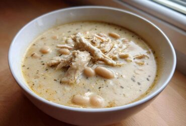 A close-up of a white bowl filled with creamy white chicken chili, featuring shredded chicken and white beans.
