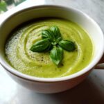 Close-up of a bowl of vibrant green zucchini soup topped with fresh basil leaves and cracked black pepper.