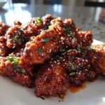 Close-up of crispy korean fried chicken recipe pieces coated in a glossy, dark sauce and topped with sesame seeds and green onions.