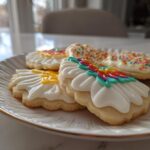 Close-up of decorated sugar cookies featuring white royal cookie icing recipe designs and sprinkles on a white plate.