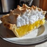A close-up of a tall slice of lemon meringue pie showing bright yellow filling, flaky crust, and peaks of toasted meringue.