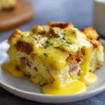 A close-up of a slice of eggs benedict casserole topped generously with rich, creamy hollandaise sauce and parsley.