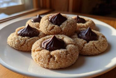 Five classic peanut butter blossom cookies, rolled in sugar and topped with a chocolate kiss, arranged on a white plate.
