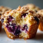 Close-up of a fluffy blueberry muffin cut in half, showing moist cake and burst blueberries, topped with streusel.