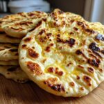 A stack of freshly cooked, fluffy homemade naan bread brushed with melted butter, showing charred spots.