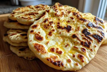 A stack of freshly cooked, fluffy homemade naan bread brushed with melted butter, showing charred spots.