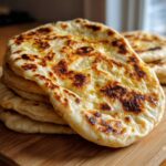 A close-up stack of freshly cooked, fluffy homemade naan bread, brushed with melted butter and showing charred spots.
