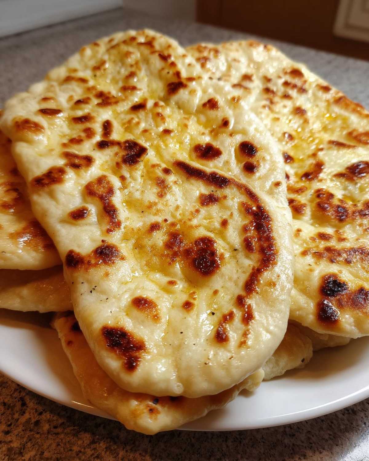 Close-up of several pieces of fluffy homemade naan bread stacked on a white plate, showing golden-brown charred spots.
