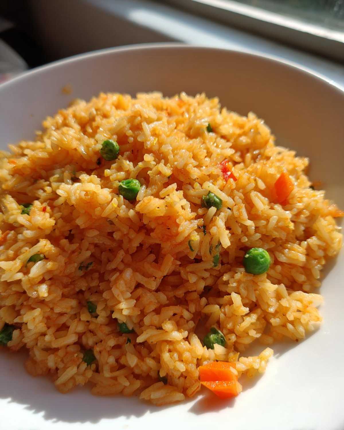 A close-up, brightly lit photo of flavorful, orange-hued spanish rice mixed with bright green peas and diced carrots in a white bowl.