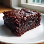 A close-up of a rich, fudgy square of vegan date brownies sitting on a white plate.