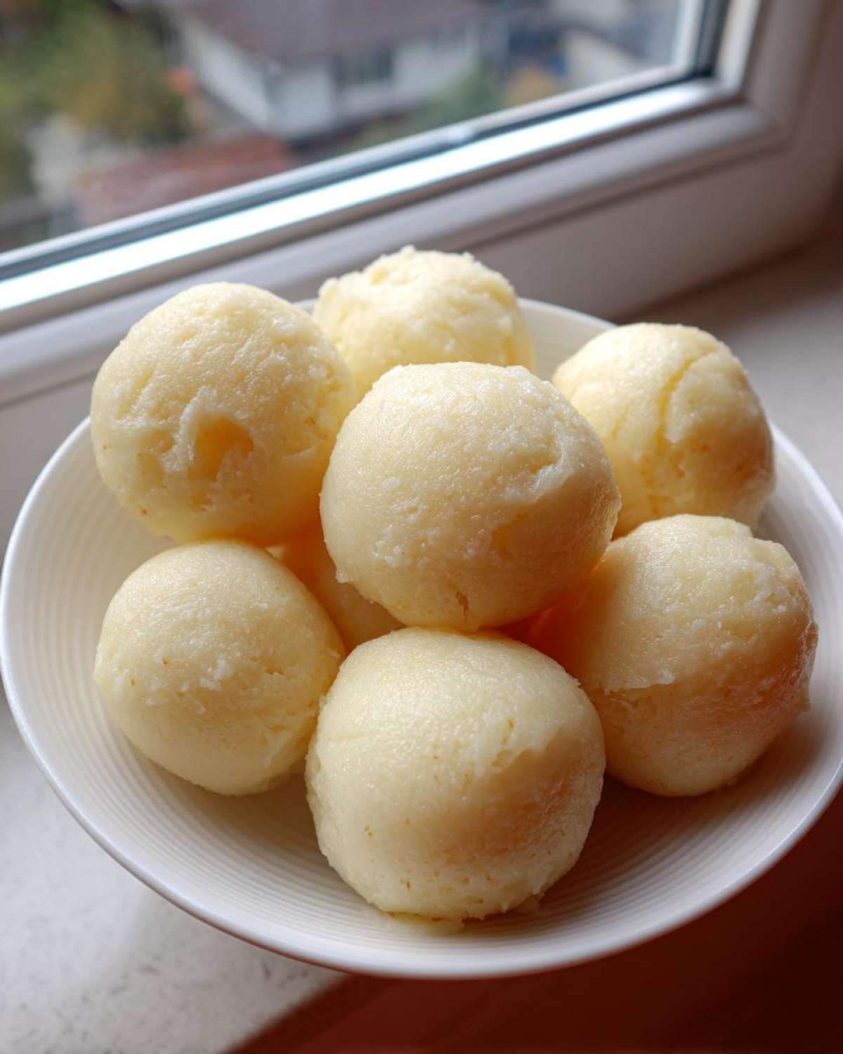 A white bowl filled with several smooth, pale yellow balls of freshly made fufu, set near a window.