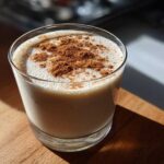 Close-up of a glass filled with creamy coquito, garnished generously with ground cinnamon on top.