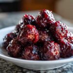 A close-up of a pile of glossy, dark red cranberry meatballs served on a white plate.