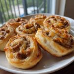 Close-up of several golden brown sausage pinwheels topped with melted cheese on a white plate.