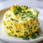A mound of bright yellow homemade garlic butter mixed with chopped green herbs, sitting on a white plate.