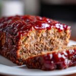 A close-up of a thick slice of juicy meatloaf topped with a glossy, dark red glaze.