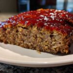 Close-up of a thick slice of juicy meatloaf topped with a shiny, thick red glaze, served on a white plate.