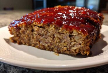 Close-up of a thick slice of juicy meatloaf topped with a shiny, thick red glaze, served on a white plate.