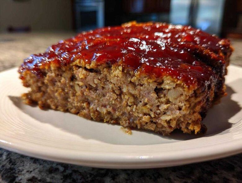 Close-up of a thick slice of juicy meatloaf topped with a shiny, thick red glaze, served on a white plate.