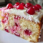 Close-up of a moist slice of vintage cherry cake, featuring a light yellow crumb studded with bright red cherries and topped with white frosting and whole cherries.