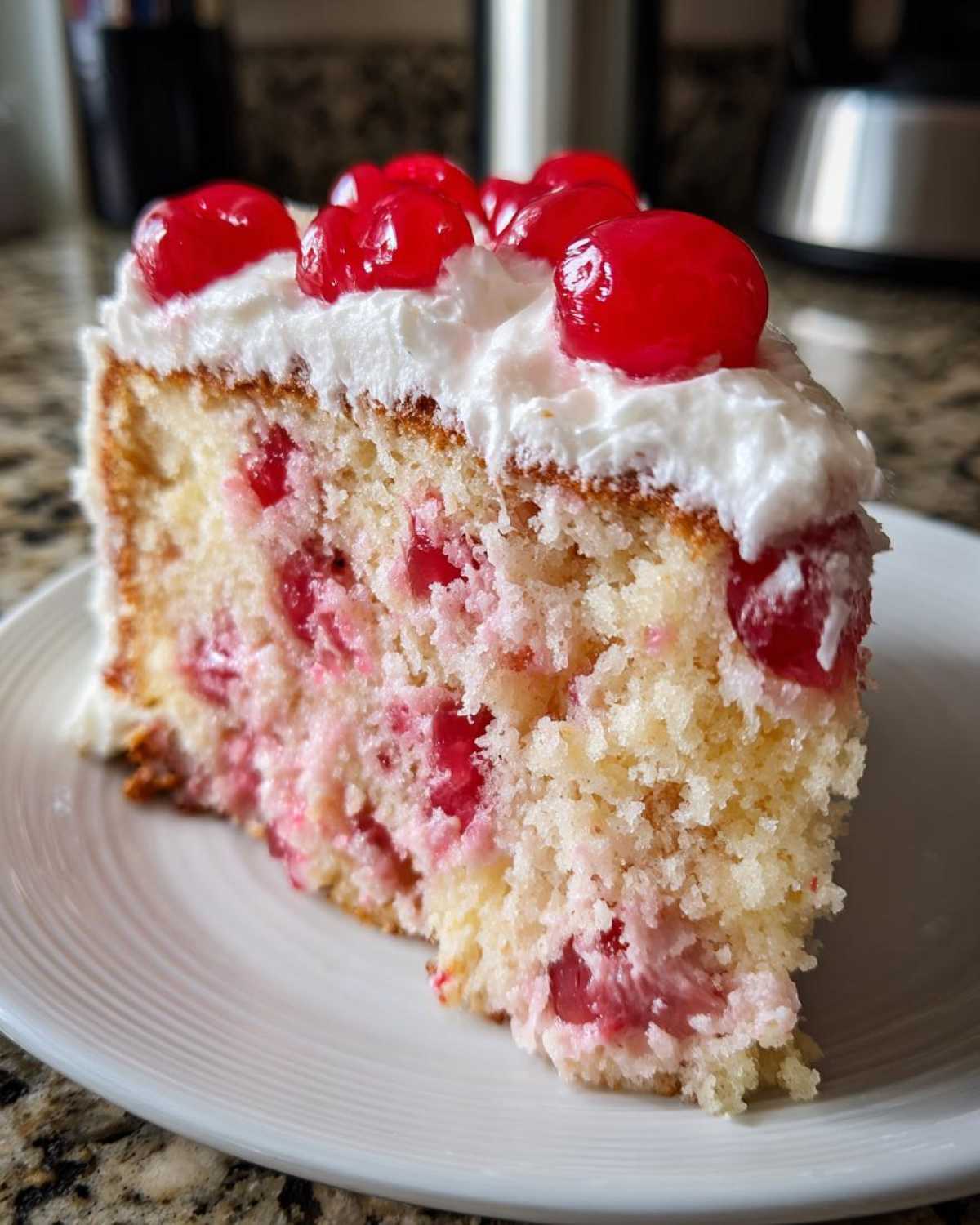 Close-up of a slice of moist cherry cake studded with pink fruit, topped with white frosting and maraschino cherries.
