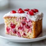 Close-up of a moist slice of vintage cherry cake, studded with red fruit and topped with white glaze and whole cherries.