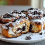Two decadent Oreo cinnamon rolls covered in white icing and crushed Oreo pieces, one cut open to show the filling.
