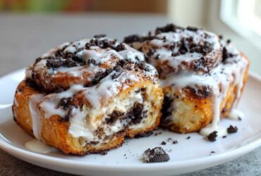 Two decadent Oreo cinnamon rolls covered in white icing and crushed Oreo pieces, one cut open to show the filling.