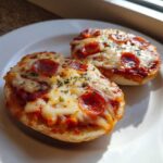 Two freshly baked pepperoni english muffin pizza halves topped with melted cheese and herbs on a white plate.