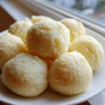 A close-up of several smooth, light yellow balls of fufu served on a white plate near a window.