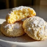 Three soft, round lemon cookies dusted heavily with powdered sugar, one broken open to show the texture.