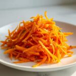 Close-up of a bright orange mound of shredded raw carrot salad seasoned with herbs and spices.