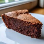 A close-up of a dense, dark chocolate slice of fudge pie dusted with cocoa powder on a white plate.