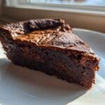 A close-up of a dense, dark chocolate fudge pie slice on a white plate, backlit by window light.