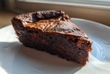 A close-up of a dense, dark chocolate fudge pie slice on a white plate, backlit by window light.