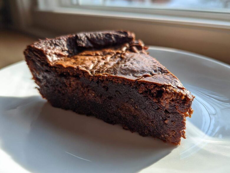 A close-up of a dense, dark chocolate fudge pie slice on a white plate, backlit by window light.