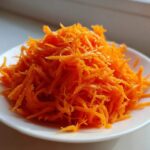 A mound of bright orange shredded carrots ready for a raw carrot salad, served on a small white plate.