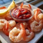 A close-up of a white platter featuring chilled shrimp cocktail pieces surrounding a small bowl of bright red cocktail sauce and lemon wedges.