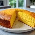A round, golden-brown skillet cornbread with one slice cut out, showing the moist interior.