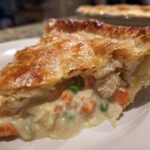 Close-up of a slice of turkey pot pie showing creamy filling, turkey, carrots, and peas under a golden, flaky crust.
