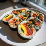 Several slices of vibrant tuna kimbap featuring rice, seaweed, tuna, egg, and greens, arranged on a white plate.