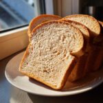 Close-up of several soft slices of homemade whole wheat bread stacked on a white plate near a sunny window.