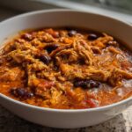 A close-up view of a hearty bowl of slow cooker buffalo chicken chili, featuring shredded chicken and kidney beans in a rich, orange-red sauce.