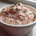A close-up view of creamy, pink smoked salmon dip topped with fresh dill, served in a white, ribbed ramekin.