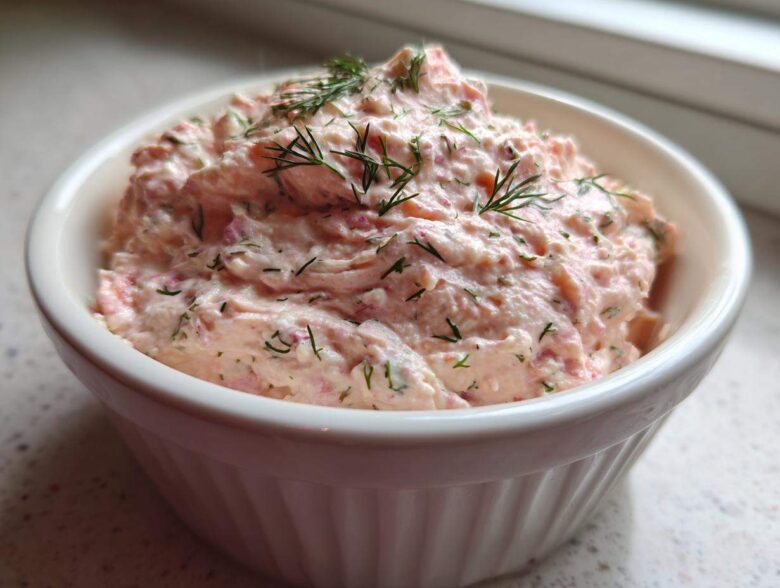 A close-up view of creamy, pink smoked salmon dip topped with fresh dill, served in a white, ribbed ramekin.
