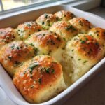 Close-up of twelve soft, buttery garlic rolls baked together in a white dish, glistening with butter and topped with fresh parsley.
