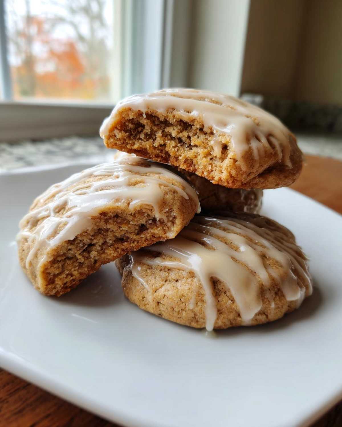Close-up of soft maple cookies stacked on a white plate, drizzled with a nutty glaze. One cookie is broken open.